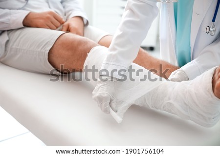 Doctor putting broken leg of young man in plaster Royalty-Free Stock Photo #1901756104
