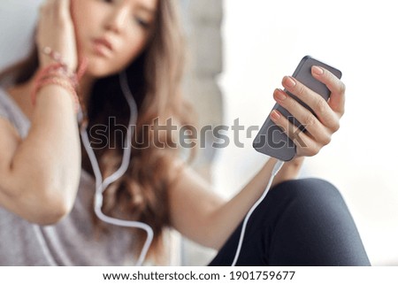 sadness and people concept - close up of teenage girl with smartphone and earphones listening to music at window