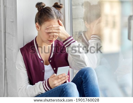 technology, cyberbullying and people concept - sad teenage girl with smartphone sitting on window sill Royalty-Free Stock Photo #1901760028