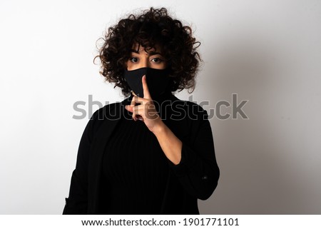 young beautiful caucasian woman wearing medical mask standing against white wall makes hush gesture, asks be quiet. Don't tell my secret or not speak too loud, please!