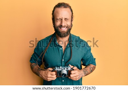 Handsome man with beard and long hair holding vintage camera smiling and laughing hard out loud because funny crazy joke. 