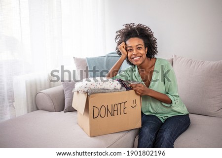 Woman putting her clothes in a carboad box with text Donate written on it. Clothes donation. Woman packing clothes into donation box in living room. Young woman with donation box at home. 