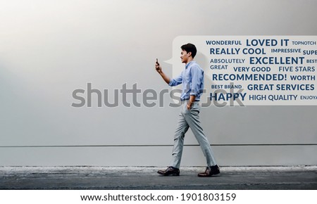 Customer Experience Concept. Reading Positive Online Review via Smartphone. Smiling Young Businessman Using Mobile Phone while Walking by the Urban Building Wall. Side View. Full Length Royalty-Free Stock Photo #1901803159
