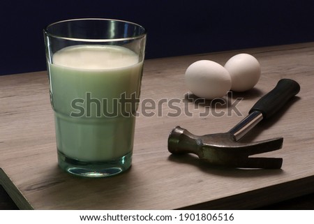 Photo joke, still life with glass of milk, two eggs and claw hammer lying on a wooden board, absurd food photo