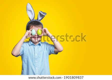 Cute boy with bunny ears keeping colored eggs near eyes on yellow background. Happy child boy with easter eggs and bunny ears on yellow background.
