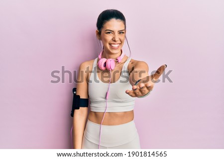 Young hispanic woman wearing gym clothes and using headphones smiling cheerful offering palm hand giving assistance and acceptance. 