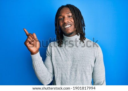 African american man with braids wearing turtleneck sweater with a big smile on face, pointing with hand and finger to the side looking at the camera. 
