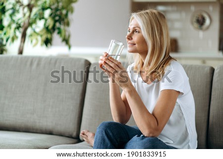Healthcare concept. Mature caucasian blonde dressed in a casual wear sits on the couch in a living room, holding a glass of pure water in a hand and smiling, follow healthy lifestyle Royalty-Free Stock Photo #1901833915