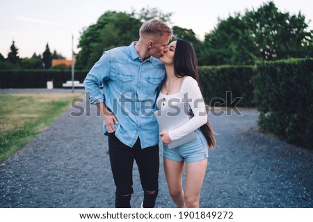 Caucasian female copywriter with digital netbook kissing boyfriend dressed in jeans shirt enjoying bonding walking date in city, good looking freelance bloggers with laptop computer hugging