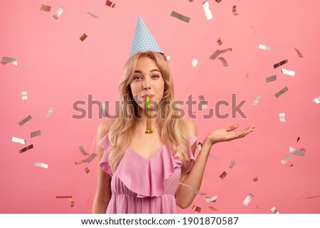 Gorgeous young woman in birthday cap blowing party horn on pink studio background with falling confetti. Positive millennial lady celebrating holiday. Festive concept Royalty-Free Stock Photo #1901870587