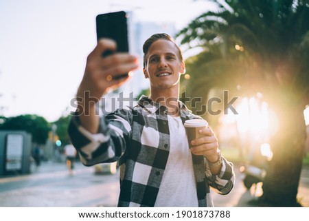 Positive young man generation z making picture of spending time in city holding coffee to go on sunny evening free time, handsome male influencer posing for selfie enjoying weekends on urban setting