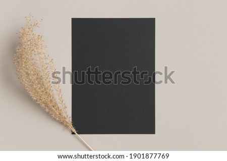 Black invitation card mockup with a dried grass decoration on a beige table. 5x7 ratio, similar to A6, A5.