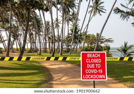''Beach closed due to COVID-19 lockdown'' information sign against a sand road amidst palms to the beach with a stretched quarantine tape