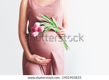 Beautiful pregnant woman with tulips flowers holds hands on belly in white background. Young woman in maternity pink dress waiting for baby birth. Pregnancy, Motherhood, Mother's Day Holiday concept. Royalty-Free Stock Photo #1901903833