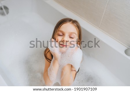 A small, smiling, beautiful red-haired girl with long hair, the child bathes, washes her face and hair on her head with foam in a white bath. Cheerful photography.
