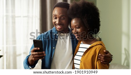 Young african american couple stands in new apartment hugging and rejoicing at receiving good news on smartphone. Concept of successful real estate purchase.