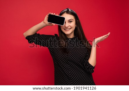 Photo of smiling beautiful brunette girl standing isolated over red wall wearing casual stylish black clothes showing mobile phone with empty screen for cutout looking at camera
