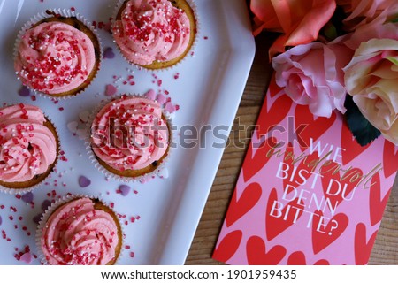 Delicious homemade pink vanilla cupcakes decorated with red hearts and roses for Valentine's day, pictured from above. The German card says "How wonderful are you please?"