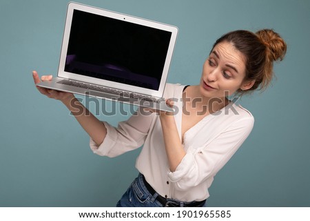 Beautiful charming young woman holding netbook computer looking down wearing white shirt isolated on blue background