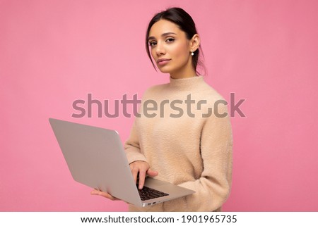 Photo of Beautiful serious young brunette woman wearing beige sweater holding netbook computer typing text on keyboard looking at camera isolated over pink wall background
