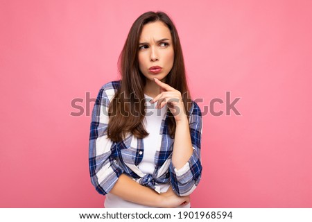 Shot of young thinking beautiful brunette woman with sincere emotions wearing trendy check shirt isolated on pink background with empty space and having doubts