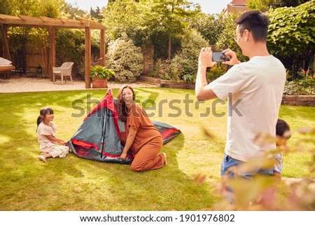 Asian Father Taking Photo On Mobile Phone As  Put Up Tent Family In Garden At Home