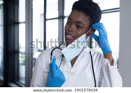 Mixed race female doctor standing putting a face mask on. medical professional healthcare worker hygiene during coronavirus covid 19 pandemic.