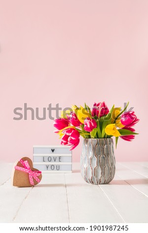 Recycled Heart shape gift box, spring bouquet of pink and yellow tulip flowers in a vase and Lightbox with love you message on the white wooden table and pink background. Vertical Greeting card mockup