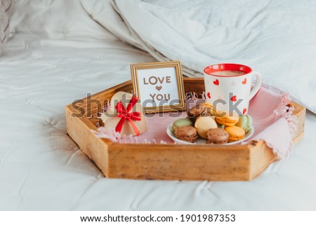 Valentine's day breakfast in bed for Lover. Love you card in the frame, a cup of coffee or cocoa, macaroons, heart-shaped gift box with red ribbon on the wooden tray. Romantic morning. Copy space.