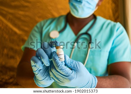 An African doctor holding a covid 19 vaccine vial in his hands while wearing blue surgical gloves and surgical scrubs Royalty-Free Stock Photo #1902015373
