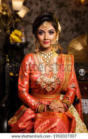 Magnificent young Indian bride in luxurious bridal costume with makeup and heavy jewellery is sitting in a chair in with classic vintage interior in studio lighting. Wedding fashion. Royalty-Free Stock Photo #1902027307