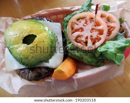 Open Swiss Cheese Hamburger with Veggies of Avocado, Lettuce, tomateos and Carrots in a red plastic basket on a table.