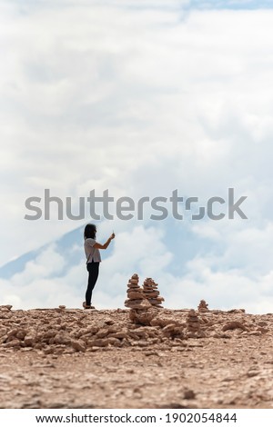 Woman taking pictures of stack of stone against cloudy sky and Lincancabur volcano in Atacama Desert, Chile