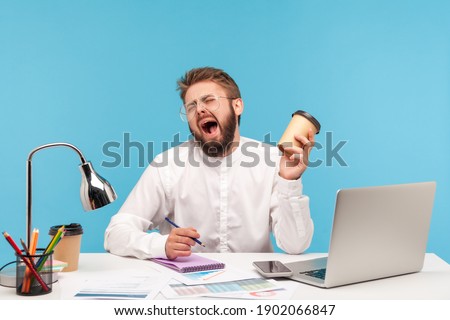 Tired man office worker with beard in eyeglasses and white shirt yawning holding paper cup of coffee and making notes in notepad sitting at workplace. Indoor studio shot isolated on blue background.