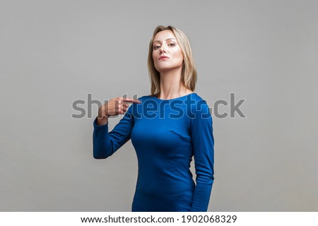 This is me! Portrait of arrogant businesswoman in tight elegant blue dress pointing at herself, looking egoistic and haughty, proud of achievement. indoor studio shot isolated on gray background Royalty-Free Stock Photo #1902068329
