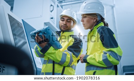 Chief Engineer and Project Manager Wearing Safety Vests and Hard Hats, Use Digital Tablet Controller in Modern Factory, Talking, Programming Machine For Productivity. Low Angle Portraits Royalty-Free Stock Photo #1902078316