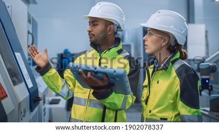 Chief Engineer and Project Manager Wearing Safety Vests and Hard Hats, Use Digital Tablet Controller in Modern Factory, Talking, Optimizing CNC Machinery, Increasing Production Line Efficiency Royalty-Free Stock Photo #1902078337