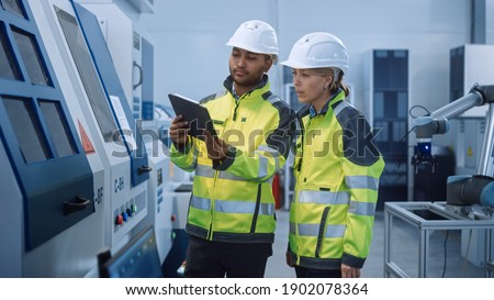 Chief Engineer and Project Manager Wearing Safety Vests and Hard Hats, Use Digital Tablet Computer in Modern Factory, Talking, Optimizing CNC Machinery, Programming Machine for Increasing Efficiency Royalty-Free Stock Photo #1902078364