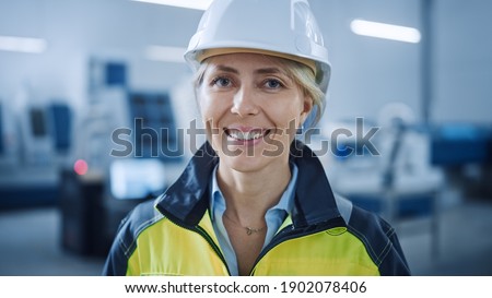 Portrait of Beautiful Smiling on Camera Female Engineer in Safety Vest and Hardhat. Professional Woman Working in the Modern Manufacturing Factory. Facility with CNC Machinery Robot arm Royalty-Free Stock Photo #1902078406