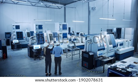 Modern Factory: Two Engineers Use Digital Tablet Computer with Augmented Reality Visualizing Workshop Room Mapping, Floor Layout. Facility with High-Tech CNC Machinery and robot arm. High Angle Royalty-Free Stock Photo #1902078631