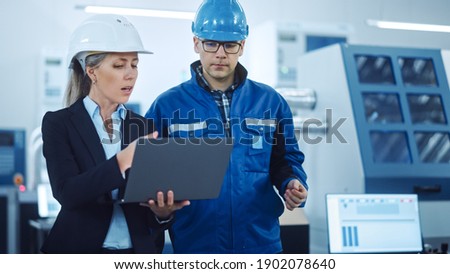 Female Manager and Project Engineer Wearing Hardhats Use Laptop in Industrial Factory, Talk, Plan Productivity Optimization. Production Line Workers Operate CNC Machinery, Program robot arm Royalty-Free Stock Photo #1902078640