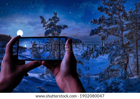 Tourist taking photo of magic winter night in a snowy sea coast surrounded by covered in snow fir trees with full moon in a starry sky. View of forest against night sky with stars and full moon.