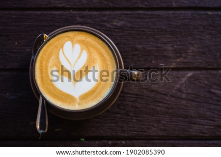 The brown beautiful ceramic cup of hot latte coffee which have latte art as a flower with spoon on the surface of the latte on the dark brown wooden table background with coyspace