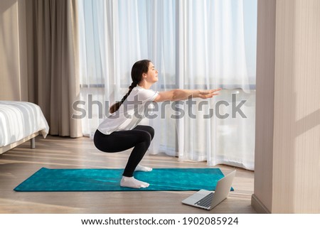A young slender woman in sportswear trains at home for online lessons. There's a laptop on the floor. Side view. Concept of home sports training on videocommunication.