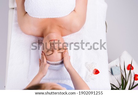 Photo from above. Hands of female masseur giving head massage to senior woman in salon with scented candles and white background.