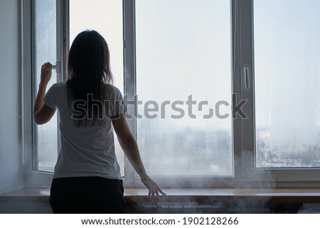 Cold winter weather. Woman step to window, open it and frosty air rushes from outside through open windows to apartment. Airing housing in cold winter. Royalty-Free Stock Photo #1902128266
