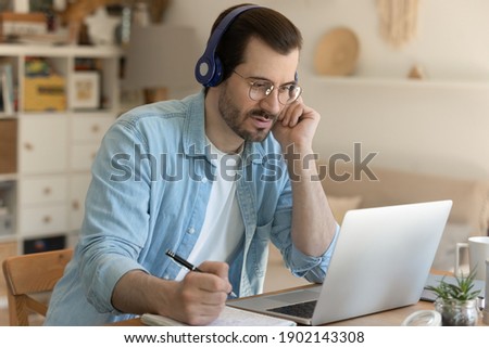 Distant studying by video conference. Focused young man student getting remote education on quarantine. Male trainee sitting in headphones by laptop listening taking notes of real time class online Royalty-Free Stock Photo #1902143308