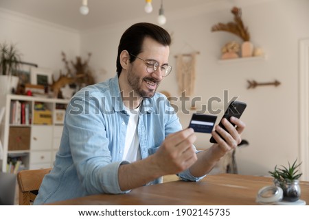 Easy paying. Happy millennial man use debit plastic card mobile telephone to provide loan mortgage payment online. Young male loyal bank client refill balance receive cash reward on electronic wallet Royalty-Free Stock Photo #1902145735