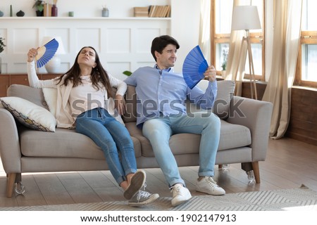 Exhausted man and woman waving blue paper fans, breathing, resting sitting on couch in living room, overheated tired couple suffering from hot summer weather, heating at home, feeling discomfort Royalty-Free Stock Photo #1902147913