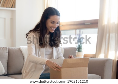Overjoyed curious young woman opening cardboard box with goods, awaited parcel at home, sitting on couch, satisfied client received online store order, good quick delivery service concept Royalty-Free Stock Photo #1902151210